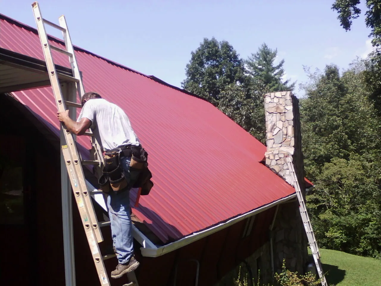 Roofer on a ladder beside a red metal roof and stone chimney — Soffit & Fascia Repair by Roofing Brothers in Cottonton
